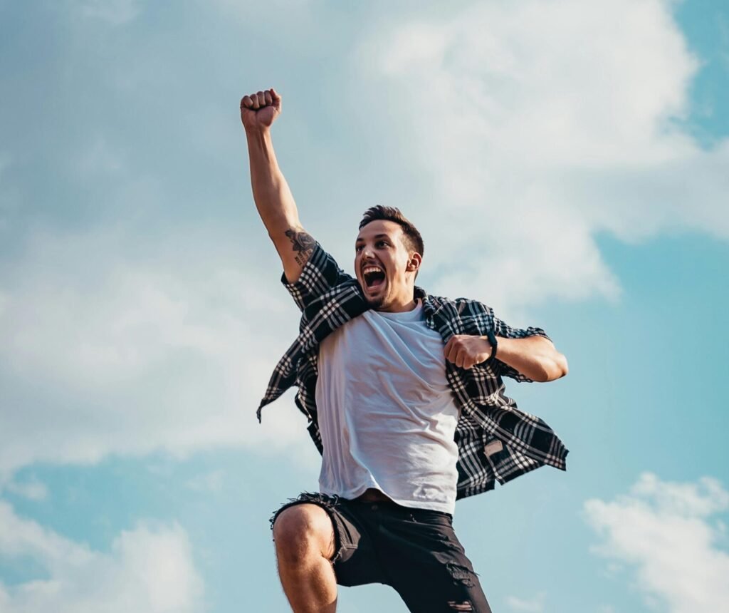 A joyful young man jumps midair with clouds and blue sky in the background, exuding energy and freedom.