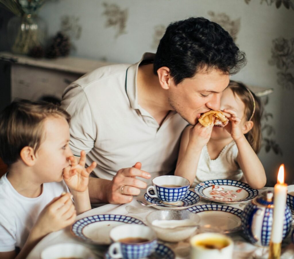 A father and his two children share a joyful meal featuring traditional crepes, creating a warm family moment indoors.